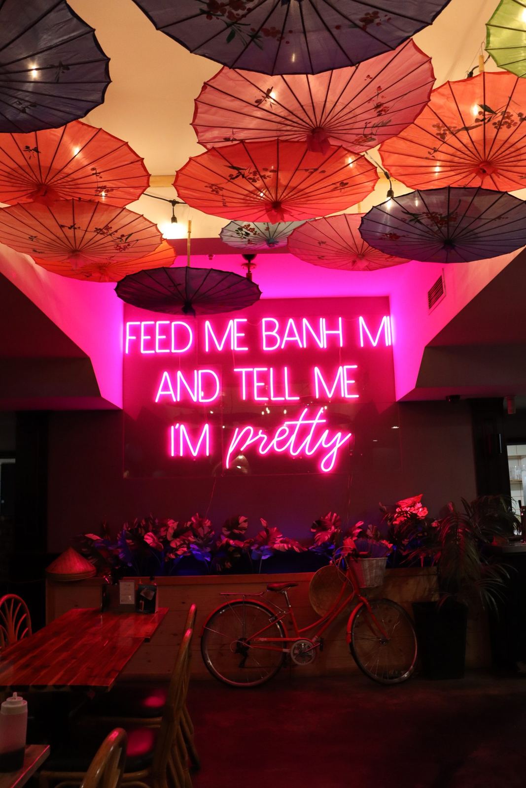 Pink neon sign under a ceiling of hanging parasols, with a vintage bicycle below